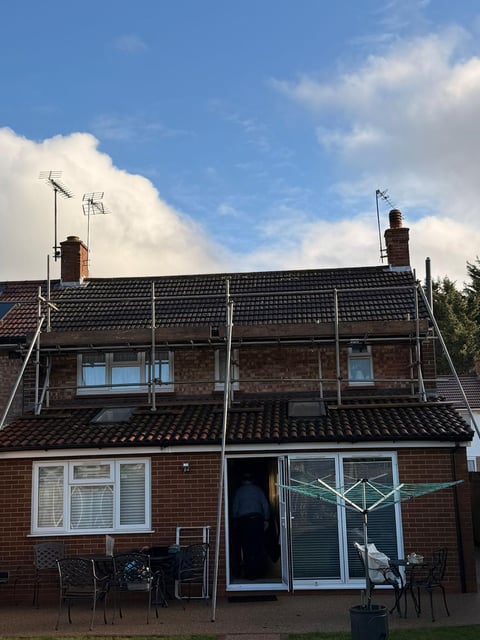 Red brick house with solar panels on roof, metal scaffolding, chimneys, and clothesline with blue sky and white clouds