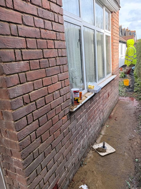 Red brick house exterior with window, worker in yellow safety vest visible in background beside narrow alley
