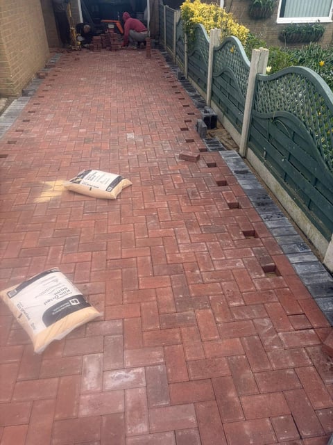 Red brick driveway with construction materials and green gates, person working in background