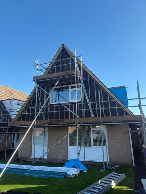Two-story residential home with steep A-frame roof undergoing renovation, surrounded by metal scaffolding under clear blue sky