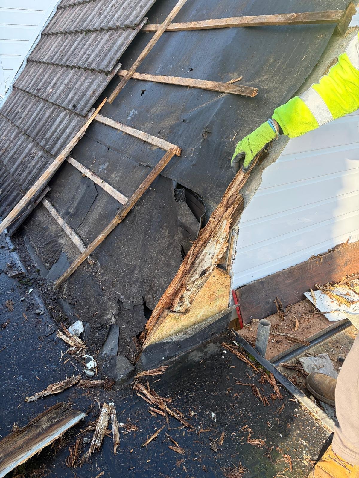 Worker in high-visibility vest inspecting deteriorated wooden beam and rusted metal roof structure during construction site assessment