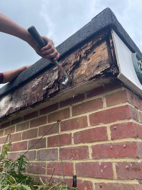 Person cleaning gutters on brick house with scraper tool, removing debris and moss buildup