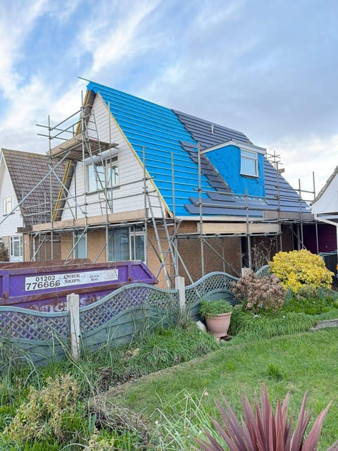 House under construction with blue roof covering and metal scaffolding, surrounded by garden landscaping