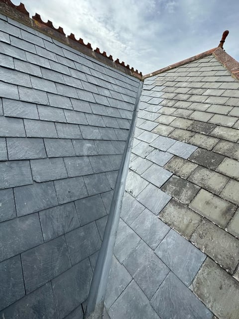 Two roof sections with slate tiles and metal gutter meeting at a ridge, showing weathered and clean shingling against a cloudy sky