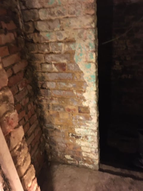 Weathered brick wall with exposed, deteriorating mortar and patches of peeling paint in an indoor space