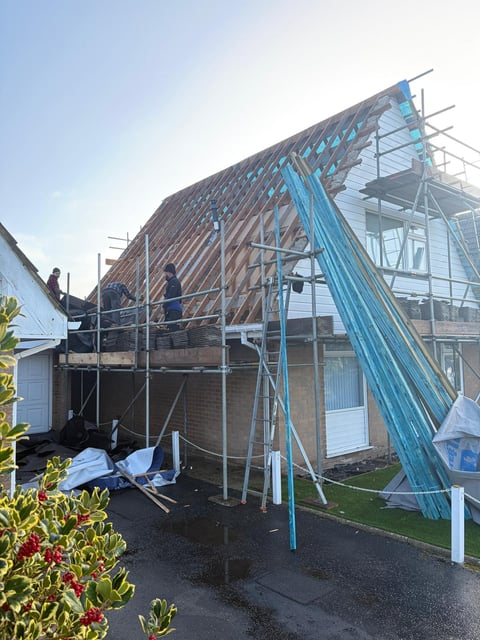 Residential house under construction with blue steel scaffolding, exposed wooden roof frame, and protective sheeting