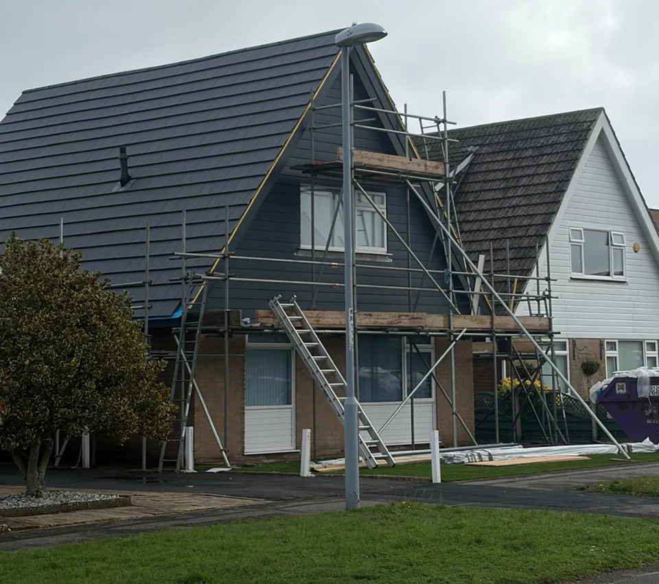 House undergoing renovation with dark roof being replaced, surrounded by metal scaffolding and construction equipment