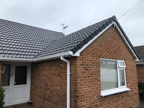 Brick bungalow with dark tiled roof, white door and window frame, and white guttering against overcast sky