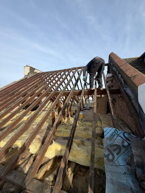 Roofer working on exposed wooden roof beams and trusses during renovation, installing new roofing materials under clear sky