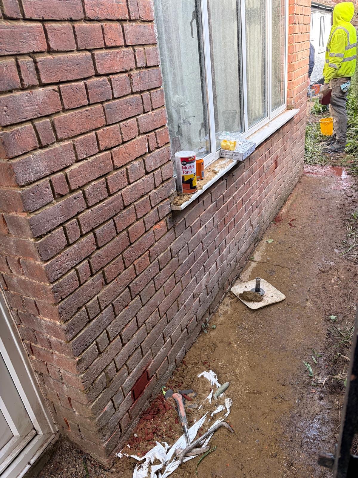 Construction site at brick building exterior with window, paint supplies on sill, and worker in yellow safety gear visible in background