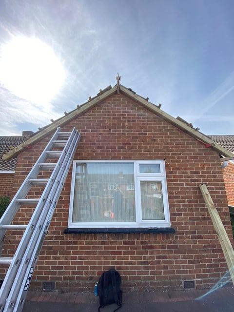 Brick house gable with aluminum ladder leaning against wall and large white-framed window, bright sunny day