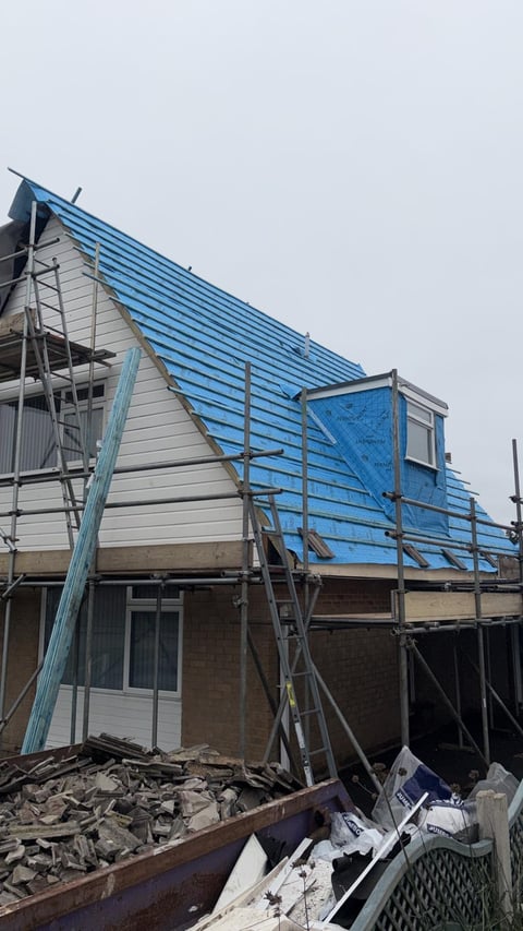 House under construction with blue metal roofing, metal scaffolding, and brick foundation surrounded by debris