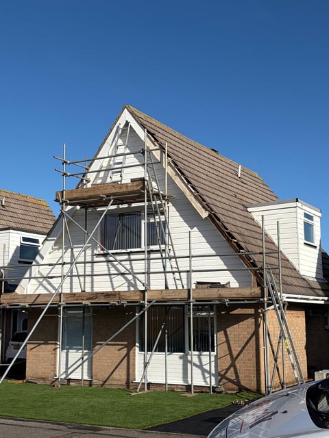 Two-story house under construction with metal scaffolding, gabled roof, and brick base against clear blue sky