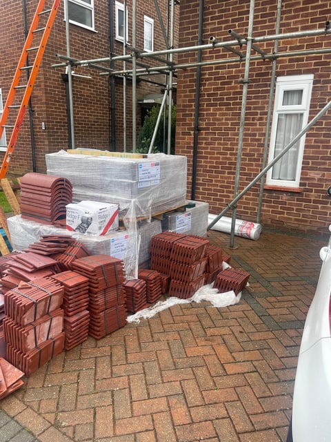 Construction site at brick building with stacks of red roof tiles and materials, metal scaffolding frame, and orange ladder