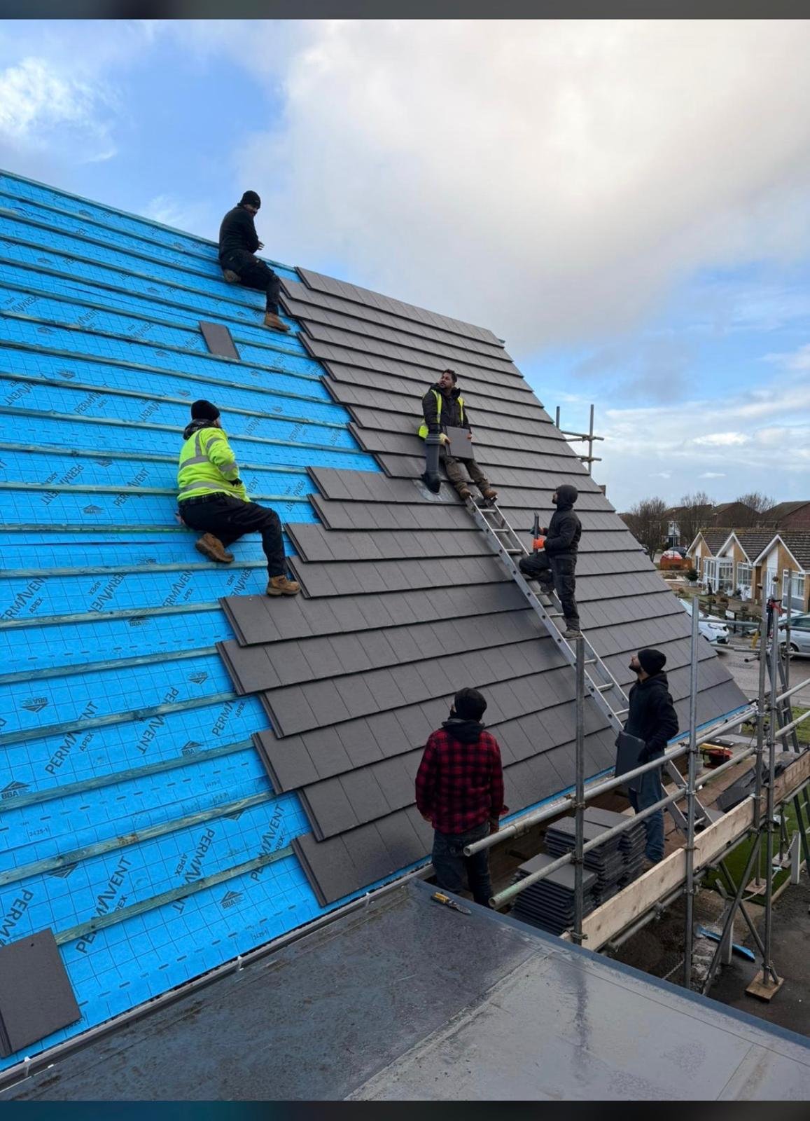 Multiple workers installing grey roof tiles on a pitched roof with blue wall sheathing, using scaffolding and safety equipment on a residential construction site