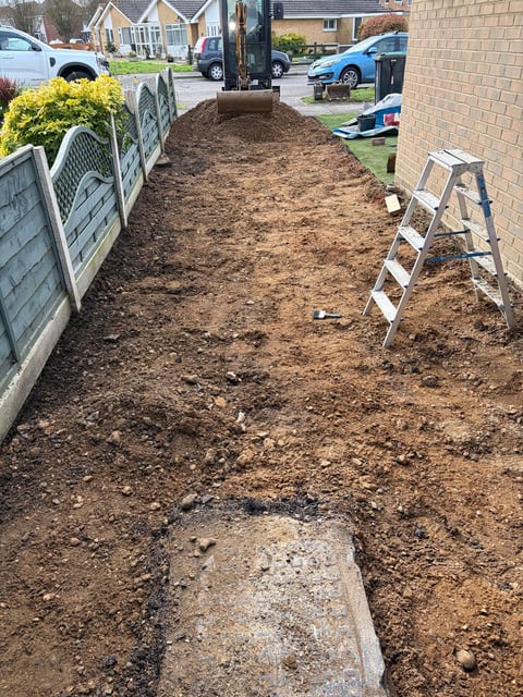 Residential driveway under construction with exposed soil, metal ladder, and white fence along the side