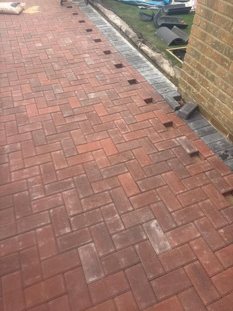 Red brick driveway with herringbone pattern extending toward a brick house wall and grassy yard