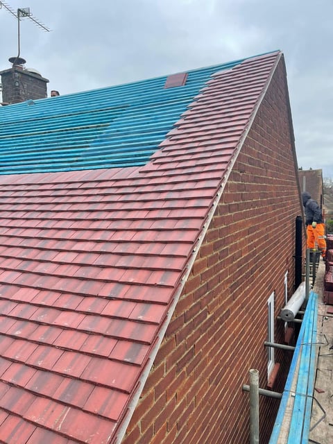Worker in orange safety gear installing blue and red corrugated metal roofing panels on a brick building under cloudy sky