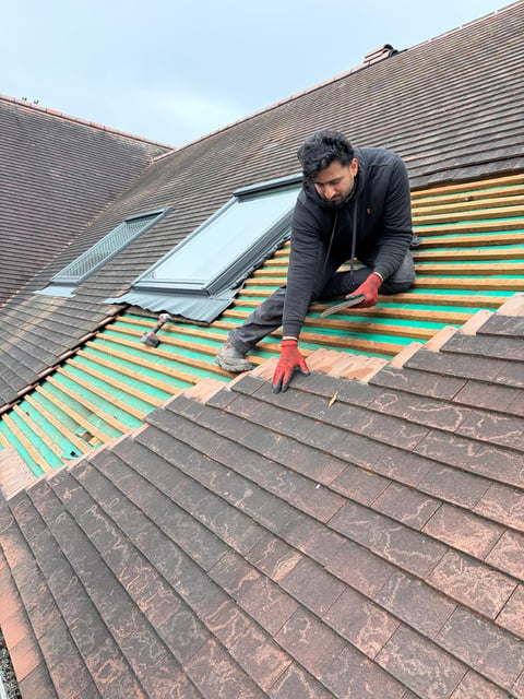 Roofer in dark jacket and red gloves installing new green roofing materials on sloped residential roof with skylights visible
