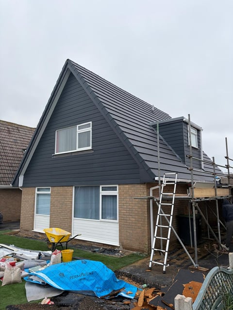 Modern house under renovation with dark blue cladding, brick base, white windows, and metal scaffolding on the right side