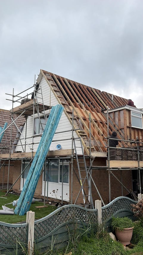House under renovation with exposed roof framing, scaffolding, and white cladding, with blue beam and garden gate visible